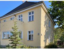 Zweistöckiges Wohnhaus mit gelber Putzfassade, weißen Fenstern und einem dunklen Schrägdach, umgeben von kleinen Bäumen und Sträuchern; eine kleine Tanne steht im Vordergrund, der Gartenbereich erstreckt sich rechts.