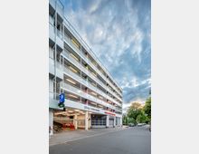 Mehrstöckiges Parkhaus mit weißer Fassade, farbigen Säulen und offenen Geländern. Im Erdgeschoss befinden sich Einfahrten mit Schildern für eine Autowaschanlage und "Herzlich Willkommen"; die Straße führt entlang eines Gehwegs mit Bäumen.