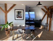 Zimmer mit offener Küche und Essbereich in einem Dachgeschoss, geprägt von sichtbaren Holzbalken. Im Vordergrund steht ein Esstisch mit grauen Stühlen, dahinter eine schwarze Küchenzeile unter Dachfenstern; Pflanzen und Dekoration sind im Raum verteilt.