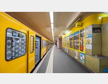 Gelber U-Bahn-Waggon an einem Bahnsteig einer U-Bahn-Station; auf der rechten Seite befindet sich ein Kiosk mit gelber Fassade und Werbeplakaten. Die Decke ist gewölbt und mit langen, hellen Leuchtstoffröhren beleuchtet.