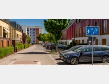 Ruhige Wohnstraße mit gepflasterter Fahrbahn, parkenden Autos entlang der rechten Seite und Bäumen in der Mitte. Links und rechts stehen moderne Reihenhäuser mit farbigen Fassaden und großen Fenstern; im Vordergrund ist ein blaues Verkehrszeichen für einen verkehrsberuhigten Bereich zu sehen.