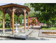Holzüberdachter Pavillon mit verziertem Dach und Marmorbrunnen in der Mitte, umgeben von einem gepflasterten Hof mit geometrischer Musterung. Im Hintergrund befinden sich eine rote Mauer, Palmen in Töpfen und Wasserbecken mit kleinen Fontänen, eingebettet in eine grüne Gartenlandschaft.