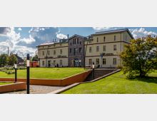 Gelbes, zweigeschossiges Gebäude mit einem verzierten Eingangsbereich und der Aufschrift "Bahnhof Zossen". Davor eine grüne Rasenfläche mit Bäumen und gepflasterten Wegen, im Hintergrund ein bewölkter Himmel.