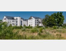 Mehrstöckige moderne Wohngebäude mit weißen Fassaden und dunklen Dächern, umgeben von einer Wiese mit niedrigem Bewuchs und vereinzelten Bäumen im Vordergrund; klare, sonnige Wetterbedingungen.