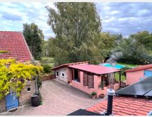 Ein ländlicher Hof mit Backsteinhäusern und roten Ziegeldächern, umgeben von Bäumen. Im Innenhof stehen ein kleines Gebäude mit Holzfenstern und ein Swimmingpool, der von Rasen und Büschen umgeben ist.