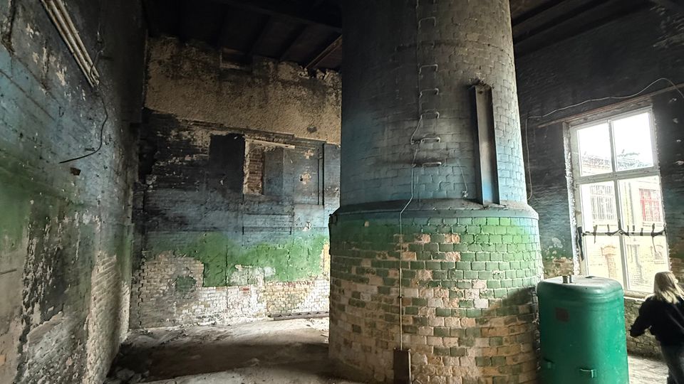Innenraum eines verlassenen Industriegebäudes mit rauen, teils abgeplatzten Ziegel- und Putzwänden und einer dunklen Holzbalkendecke. In der Mitte steht ein großer zylindrischer gemauerter Ofen oder Schornstein, daneben ein grüner Tank; rechts fällt Licht durch ein großes Fenster.