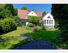 Zweistöckiges Einfamilienhaus mit weißer Fassade und roten Dachziegeln, umgeben von einem Garten mit gepflegtem Rasen, Bäumen und bepflanzten Flächen. Ein gepflasterter Weg führt zu einer Terrasse mit Sitzmöbeln, umrankt von Kletterpflanzen.