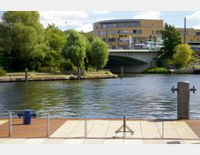 Uferbereich mit Pflastersteinen und einer Metallabsperrung im Vordergrund, dahinter ein Fluss mit Bäumen und einer Anlegestelle. Im Hintergrund eine moderne Brücke und ein Gebäude aus gelbem Backstein mit Geschäften, darunter ein Netto-Markt.
