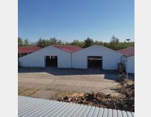 Mehrere weiße Hallengebäude mit dunkelroten Dächern stehen in einer betonierten Freifläche, umgeben von Bäumen unter klarem, blauem Himmel. Im Vordergrund sind Holz- und Ziegelreste neben einem Wellblechdach zu sehen.