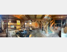 Innenraum eines rustikalen Scheunen- oder Dachbodenraums mit freiliegenden Holzbalken, Backsteinboden und steinernen Wänden, teils als Atelier mit Staffelei und gerahmten Gemälden genutzt. Rechts führt eine Holztreppe mit Geländer zu einer Galerieebene; im Vordergrund stehen ein runder Tisch mit Stühlen sowie eine Couch.