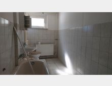 Ein einfaches Badezimmer mit weiß gefliesten Wänden und Boden. Es enthält eine Badewanne, ein Waschbecken, eine Toilette und einen Heizkörper unter einem kleinen Fenster.