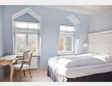 Helles Schlafzimmer mit zwei großen Fenstern, die von Dachschrägen eingerahmt sind. Im Raum befinden sich ein Doppelbett mit weißer Bettwäsche, eine Holzkommode mit Stuhl sowie ein heller Holzboden und pastellblaue Wände.