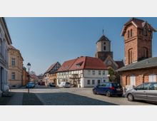 Straßenszene in einer historischen Altstadt mit Kopfsteinpflaster, umgeben von Fachwerkhäusern und Backsteinbauten. Im Hintergrund ragt ein Kirchturm hervor, und geparkte Autos sind entlang der Straße zu sehen.