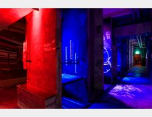 Ein dunkel beleuchteter Flur mit Industriedesign, Wänden aus Beton und sichtbaren Rohren an der Decke. Die Szene ist in rotes und blaues Licht getaucht, mit Graffiti und Nummerierungen ("Zelle 17-19") an den Wänden.