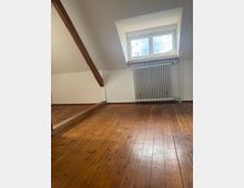 Leerer Dachraum mit schrägen weißen Wänden, sichtbaren Holzbalken und breiten Dielen-Holzboden. An der Stirnseite befindet sich ein kleines Dachfenster mit Heizkörper darunter.