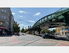 Straßenkreuzung in einer Stadt mit Straßenbahnschienen im Vordergrund, flankiert von mehrstöckigen Wohnhäusern links und rechts. Über der Straße verläuft eine grüne Hochbahnbrücke, im Hintergrund sind der Fernsehturm und eine Kirchturmspitze sichtbar.
