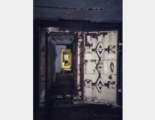 Blick in einen schmalen, betonierten Gang mit einer geöffneten, schweren Stahltür mit rostigen Beschlägen und Verriegelungsmechanismus, typisch für einen Bunker oder Schutzraum. Im Hintergrund führt der Korridor durch weitere Türrahmen zu einem helleren Raum.