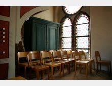 Innenraum einer Kirche mit einer Reihe von Holzstühlen, einem großen, grünen Schrank vor der Wand und zwei hohen Fenstern mit farbigem Buntglas in Spitzbogenform. Links ist ein Teil einer Orgel mit Knöpfen zu sehen.