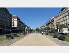 Breite Straßenansicht in einer Stadt mit symmetrischen Gebäuden mit klassischen Fassaden auf beiden Seiten, in der Mitte ein Fußweg mit gepflastertem Rand, Bänken und Fahrrädern. Im Zentrum ist ein U-Bahn-Eingang mit einem blauen "U"-Schild, umgeben von Grünflächen und Bäumen.