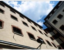 Blick auf die Fassade eines mehrstöckigen historischen Backsteingebäudes mit verputzten Wänden und Fenstern, die durch Ziegelsteinrahmen eingefasst sind und teils Gitter aufweisen. Im Vordergrund ist eine Überwachungskamera montiert, während im Hintergrund blauer Himmel mit Wolken sichtbar ist.