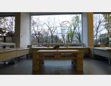 Ein moderner Loungebereich mit hellen Polstermöbeln und einem niedrigen Tisch aus Holzpaletten vor einer großen Fensterfront. Draußen sind kahle Bäume und Gebäude in einer urbanen Umgebung zu sehen, mit einer dekorativen Zeichnung auf der Fensterscheibe.