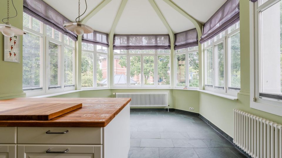 Heller Wintergarten-ähnlicher Raum mit vielen bodentiefen Fenstern rundum, hellgrünen Wänden und sichtbaren Deckenstreben unter einem spitz zulaufenden Dach. Vorne steht eine Kücheninsel mit Holzarbeitsplatte; entlang der Wände sind Heizkörper und graue Fliesen am Boden.
