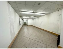 Ein leerer, langer Flur mit weißen Wänden, deckenintegrierter Beleuchtung und einem gefliesten Boden. Am Ende des Flures befindet sich eine geschlossene Metalltür, die möglicherweise zu einem Aufzug gehört.