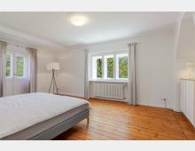 Helles Schlafzimmer mit weiß gestrichenen Wänden, Holzdielenboden und großem Fenster mit Vorhängen sowie Heizkörper darunter. Im Raum stehen ein Bett, eine Stehlampe und ein Sideboard; links ist eine verglaste Tür mit Vorhängen sichtbar.