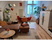 Ein gemütliches Wohnzimmer mit Holzfußboden, einem braunen Ledersofa, einem Couchtisch mit Blumen, und einer modernen Stehleuchte mit bunten Schirmen. Im Hintergrund steht ein runder Esstisch mit farbigen Stühlen vor einem großen Fenster, das auf einen Balkon mit Blumen blickt; daneben ein weißer Schrank und eine Gitarre an der Wand.