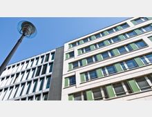 Moderne Bürogebäude mit mehrstöckiger Fassade, gestaltet mit großen Fenstern und grünen Paneelen, neben einem grauen Abschnitt mit vertikalen Fensterelementen. Im Vordergrund steht eine Straßenlaterne, der Himmel ist klar und blau.