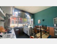 Innenansicht eines kleinen Restaurants oder Imbisses mit türkischem Dekor. Im Vordergrund befinden sich eine Theke mit Küchengeräten und ein aufgestellter Dönerspieß, während durch große Fenster der Blick auf eine Straße mit Backsteingebäuden fällt.