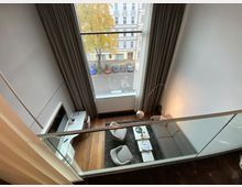 Moderne Maisonette-Wohnung mit Blick in den Wohnbereich, der ein großes Fenster bis zur Decke, graue Vorhänge, eine graue Sofalandschaft, zwei Sessel und einen Couchtisch umfasst. Im Vordergrund befindet sich eine Glasbrüstung; außen ist eine Straßenansicht mit parkenden Autos und Bäumen sichtbar.