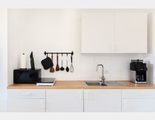 Schlichte Küche mit weißem Schrank und Holzarbeitstisch, ausgestattet mit einem Spülbecken, Kaffeemaschine, Mikrowelle und induktiver Kochplatte. An einer schwarzen Stange sind Kochutensilien und ein Topflappen aufgehängt.