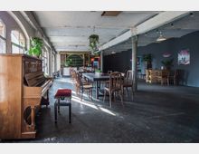 Großzügiger Raum im Industriestil mit unverputzten Wänden und Deckenbalken. Im Vordergrund steht ein hölzernes Klavier, daneben mehrere Holztische und Stühle sowie Pflanzen zur Dekoration. Im Hintergrund ist eine Theke mit Regal und weiteren Sitzgelegenheiten sichtbar.