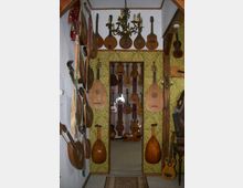 Schmaler Raum mit gelb-grüner Tapete und zahlreichen Saiteninstrumenten, darunter Gitarren, Lauten und Mandolinen, die an den Wänden hängen. In der Mitte des Raumes hängt ein großer, verzierter Kronleuchter aus Metall, und durch einen Türrahmen ist ein weiterer Raum mit zusätzlichen Instrumenten sichtbar.