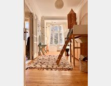 Ein helles Kinderzimmer mit hohen Decken und verzierten Stuckelementen. Das Zimmer enthält ein Hochbett mit einer Leiter, ein kleines Schreibtisch-Set, große Fenster mit weißen Vorhängen und einen gemusterten Teppich auf Dielenboden.