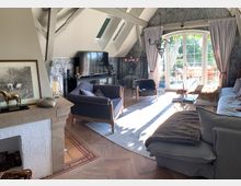 Wohnzimmer mit Dachschrägen, einem großen Panoramafenster mit bodenlangen Vorhängen und viel Tageslicht. Die Einrichtung besteht aus einem hellen Sofa, blauen Sesseln, einem Kamin, einem Fernseher und einem Glastisch, ergänzt durch Holzboden und dekorative Akzente wie ein Bild und Leuchter.