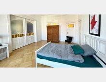 Helles Schlafzimmer mit einem Holzboden, einem weißen Bett mit grauer Bettwäsche, einem hölzernen Kleiderschrank und weißen Türen mit gelben Glaseinsätzen. An der Wand hängt ein gerahmtes Poster, und in einer Ecke steht ein schwarzer Sessel.