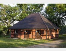 Fachwerk-Pferdestall mit Backsteinmauern und dunklem Satteldach, umgeben von grünen Bäumen und Wiesen; mehrere Pferde schauen aus den offenen Stallfenstern.