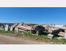 Ein offenes Lagergelände mit Betonplatten und -trümmern, die in Stapeln auf unebenem, bewachsenen Boden liegen. Im Hintergrund sind Holzhaufen, runde weiße Folienballen und ein bewaldeter Horizont bei klarem, blauem Himmel zu sehen.