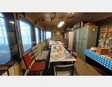 Ein einfacher Raum in einem alten Zugabteil mit Metallspinden, einer langen Tischgruppe mit Stühlen, und verschiedenen Gegenständen wie Zeitungen und Geschirr. Die Fenster auf der linken Seite lassen Licht durch und geben den Blick auf ein Gittermuster im Außenbereich frei.