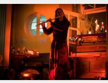 Eine Person spielt eine Querflöte in einem warm beleuchteten Raum mit einer gelben Wand und einem runden Fenster. Im Hintergrund sind ein Klavier, Kerzen und verschiedene Dekorationsgegenstände sichtbar.