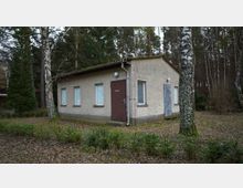 Kleines, rechteckiges Gebäude mit hellgrauer Fassade und flachem Dach, ausgestattet mit mehreren Fenstern und einer braunen Tür, auf der „Dusche“ steht; umgeben von Laubbäumen und niedrigem Pflanzenbewuchs in einer waldähnlichen Umgebung.