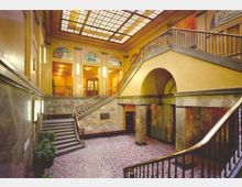 Historisches Treppenhaus mit gelben Wänden, Marmorsäulen und einem farbigen Glasfenster als Decke. Die Treppe führt zu einer oberen Etage mit Gewölbebögen und Wandmalereien.