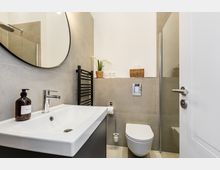 Modernes Badezimmer mit grauen Fliesen, weißem Waschtisch mit rundem Spiegel und wandhängendem WC. Rechts ist eine Glasdusche zu sehen, dazu ein schwarzer Handtuchheizkörper und kleine Körbe sowie eine Pflanze als Ablageelemente.