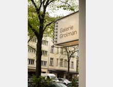 Straßenszene mit einem Schild, auf dem „Galerie Grolman“ steht, angebracht an einem Gebäude neben Laubbäumen. Im Hintergrund sind eine belebte Straße, geparkte Autos und ein mehrstöckiges Wohngebäude mit großen Fenstern zu sehen.