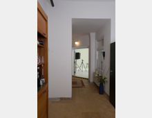 Ein schmaler Flur mit gelbem Fliesenboden, links ein Holzschrank mit Flaschen und Gläsern, rechts eine blaue Vase mit Pflanzen. Im Hintergrund ein heller Raum mit einem Kamerastativ und Hockern auf einem weißen Boden, getrennt durch eine Schwelle mit gelb-schwarzer Markierung.