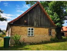 Altes Landhaus aus Backstein mit einem Giebeldach, gedeckt mit roten Ziegeln. Die obere Hälfte der Fassade ist aus dunklem Holz gefertigt, umgeben von Bäumen und einer Rasenfläche; ein grüner Regentonnenbehälter steht links im Vordergrund.