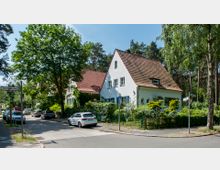 Straßenszene in einer ruhigen Wohngegend mit Einfamilienhäusern im traditionellen Stil, weißen Fassaden und roten Ziegeldächern. Im Vordergrund stehen parkende Autos, daneben hohe Bäume und ein Gehweg; im Hintergrund ist ein Wald erkennbar.