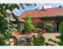 Hof mit Backsteingebäude und roten Ziegeldächern, umgeben von Pflanzen. Im Innenhof stehen Holztische und Stühle sowie Blumendekorationen, mit direktem Zugang zu großen grünen Doppeltoren und einem hellen Anbau mit Fenstern.